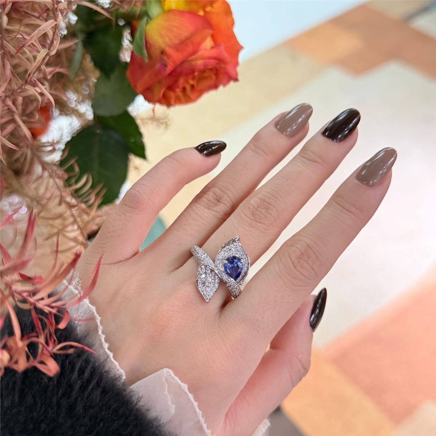 A close-up of a hand wearing a silver ring with a central blue teardrop-shaped gemstone and smaller clear stones around it. The hand has brown manicured nails, and a blurred orange rose and warm-toned background are visible.